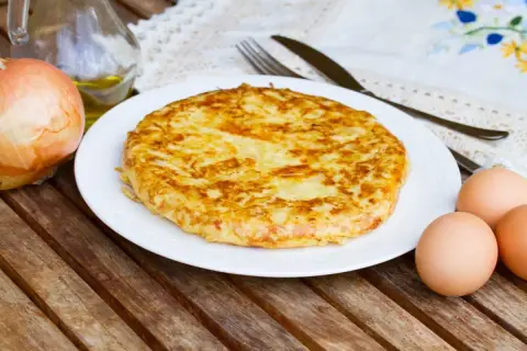 Frittata di patate dorata su piatto bianco, con cipolla, uova e olio d'oliva come ingredienti.